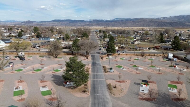 Aerial view of an RV park with empty parking spaces, small trees, and a road running through a residential area with mountains in the background.