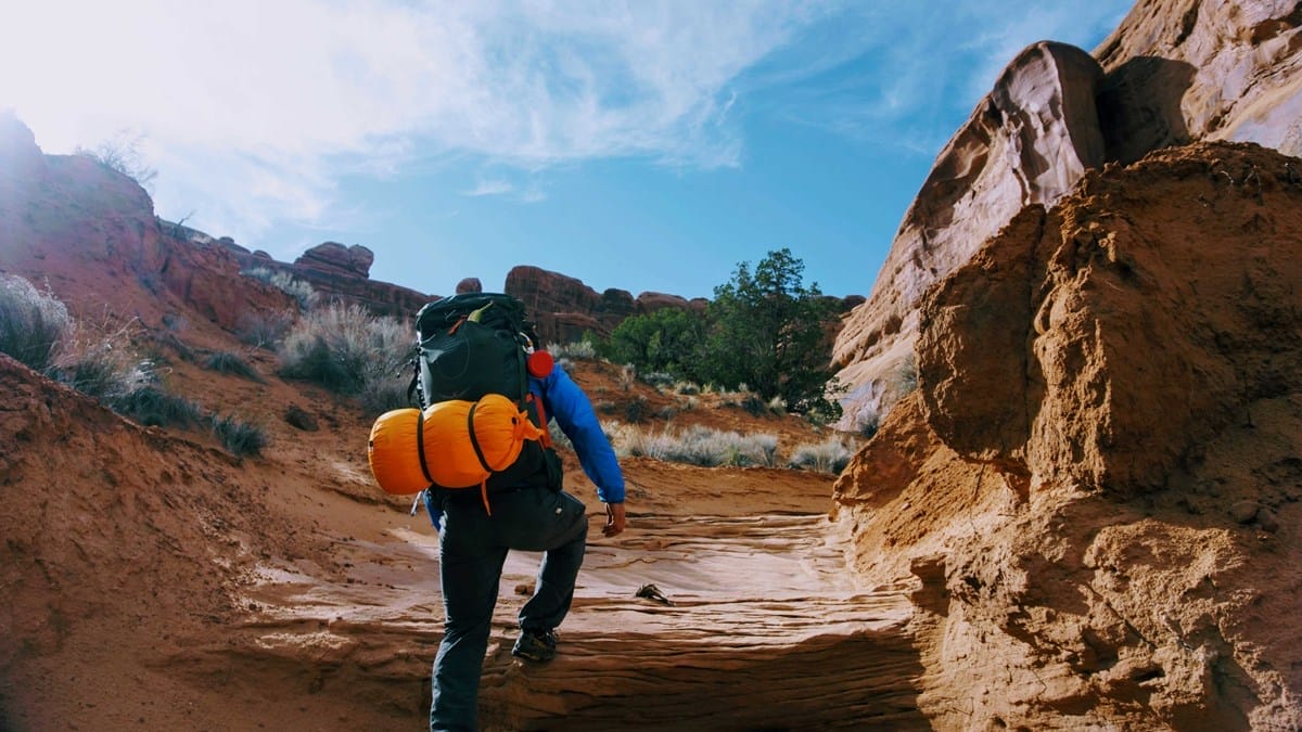A person with a large backpack and sleeping pad hikes up a rocky, sandy trail surrounded by cliffs and sparse vegetation under a blue sky.