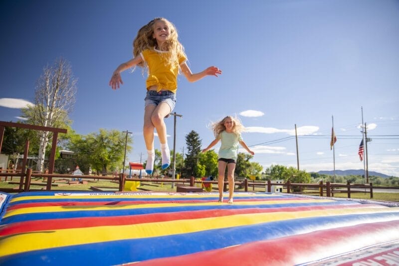 Two children jump on a large, colorful inflatable mat outdoors on a sunny day with trees, flags, and a blue sky in the background.