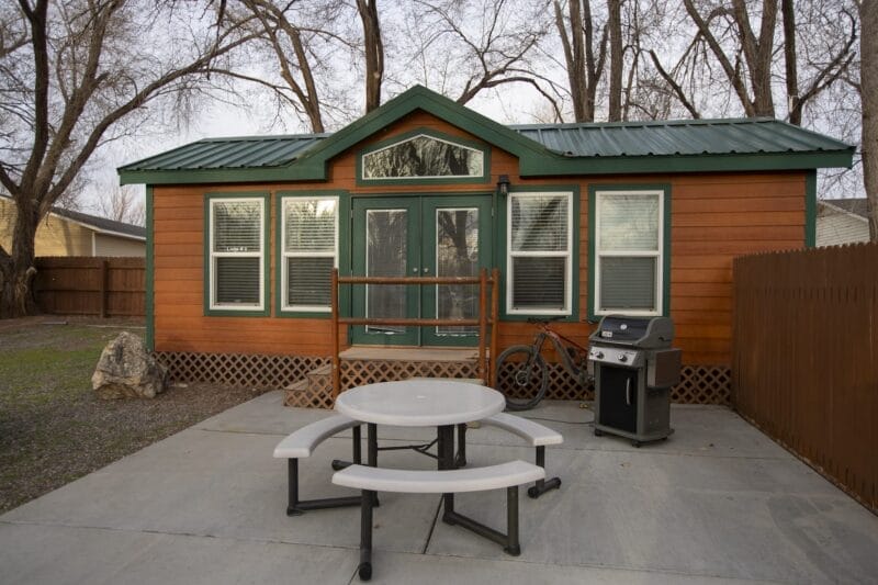 Small wooden cabin with green trim, three windows, glass double doors, a round picnic table, a grill, a wooden rack, and a bicycle on a concrete patio. Trees and a fence are in the background.