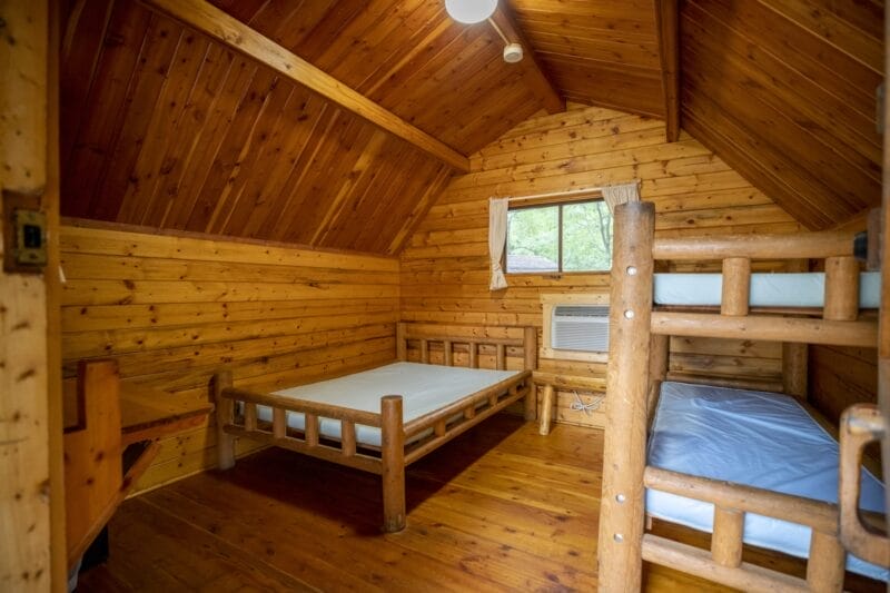 A wooden cabin room with a single bed, bunk beds, small window, air conditioning unit, and wooden walls, floor, and ceiling.