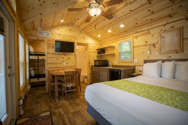 A small wooden cabin interior with a bed, bunk beds, dining table, kitchenette, TV, ceiling fan, and bright lighting.