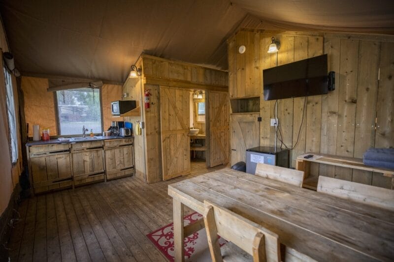 Interior of a rustic wooden cabin with a dining table, chairs, kitchen area, wall-mounted TV, and visible bathroom entrance.