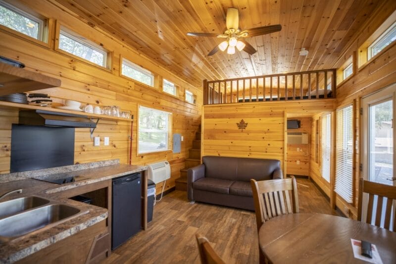 Interior of a small wooden cabin with a kitchenette, sofa, dining table, and a loft area; ceiling fan and windows provide natural light.