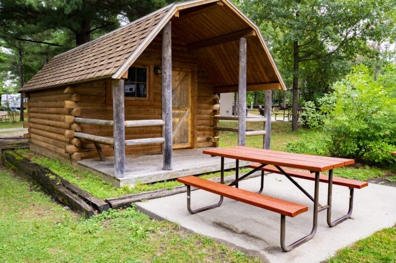 A small wooden cabin with a covered porch sits beside a red picnic table on a concrete pad, surrounded by grass and trees.
