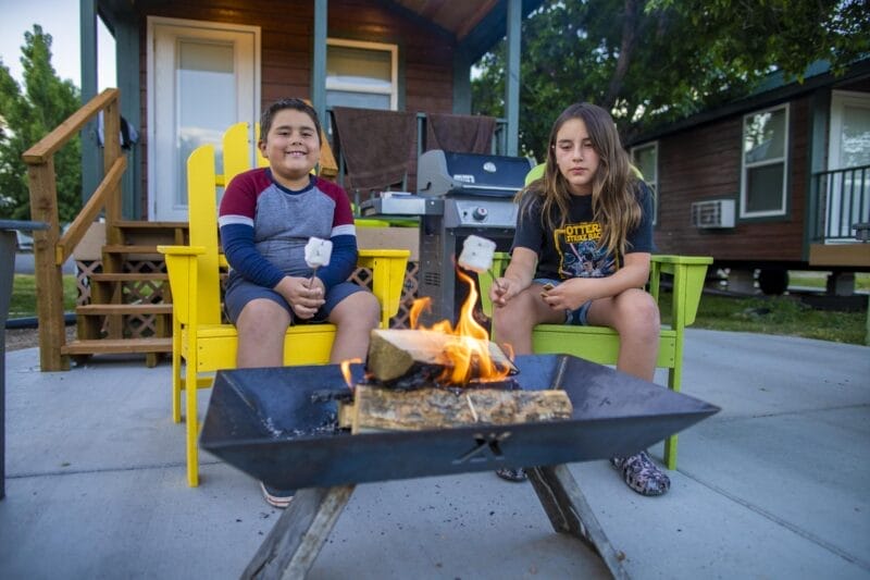 Two children sit in yellow chairs outside a cabin, roasting marshmallows over a small fire pit on a patio.