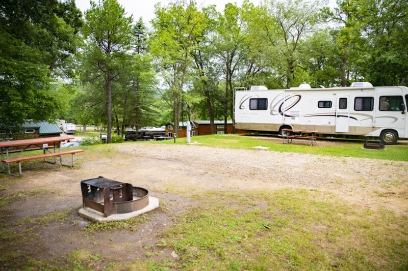 A campsite with a gravel pad, fire pit, picnic table, and a parked RV surrounded by trees and cabins in the background.