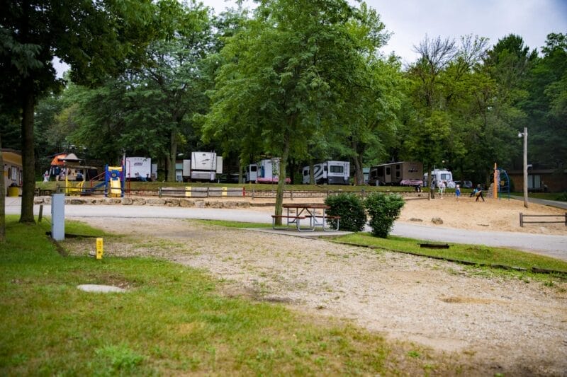 Several RVs are parked near a sandy playground area surrounded by trees at a campground, with a picnic table and grassy area in the foreground.