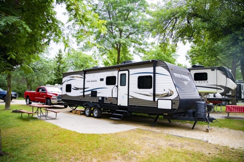 A white and gray travel trailer is parked at a campsite beside a red pickup truck, with trees, picnic tables, and other RVs visible nearby.