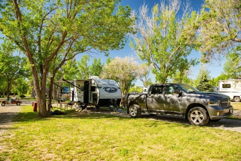 A black pickup truck is parked beside a white camper trailer at a shaded campground with green grass and trees under a clear blue sky.