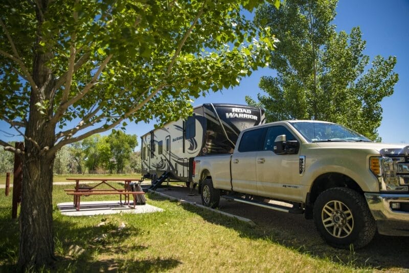 A pickup truck towing a large Road Warrior RV is parked beside a picnic table under a leafy tree in a grassy, sunny campground.