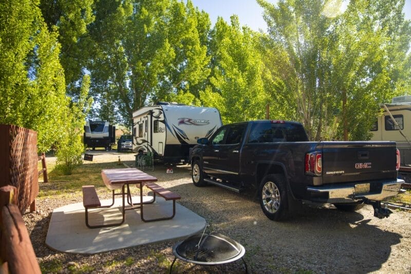 A black GMC pickup truck is parked beside a travel trailer at a campsite with a picnic table, surrounded by trees.