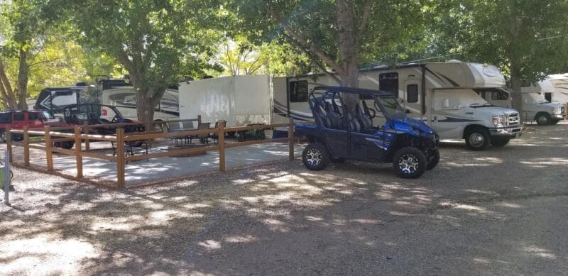 Several RVs and a side-by-side utility vehicle are parked under trees at a shaded campsite with a fenced seating area.