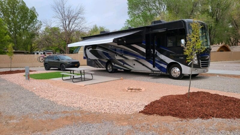 A large RV with an extended awning is parked next to a black car at a campsite with a picnic table and small tree, surrounded by gravel and mulch landscaping.