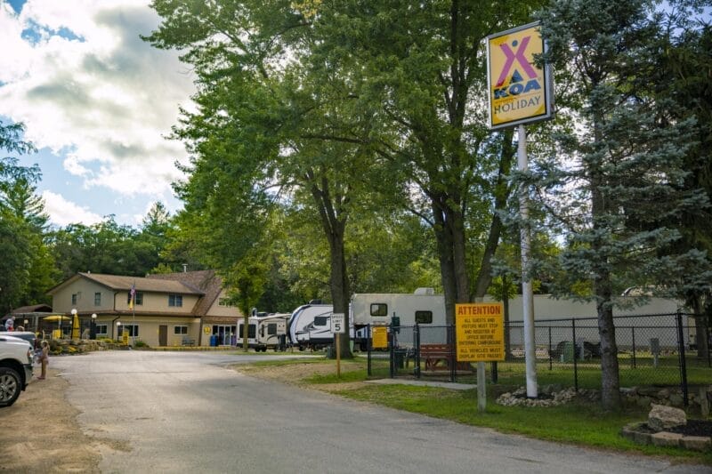 A KOA Holiday campground entrance with a sign, parked RVs, a building, and trees on a partly cloudy day.