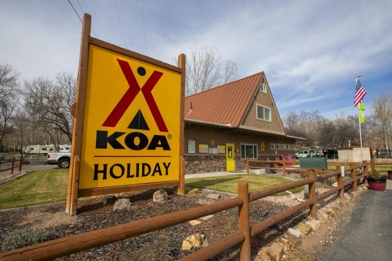 A large KOA Holiday campground sign stands in front of an office building, with RVs, trees, and flags visible in the background.