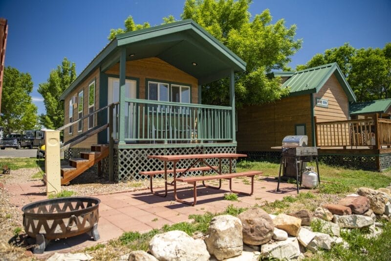 Small cabin with a front porch, picnic table, grill, and fire pit in front, surrounded by rocks and trees under a clear blue sky.