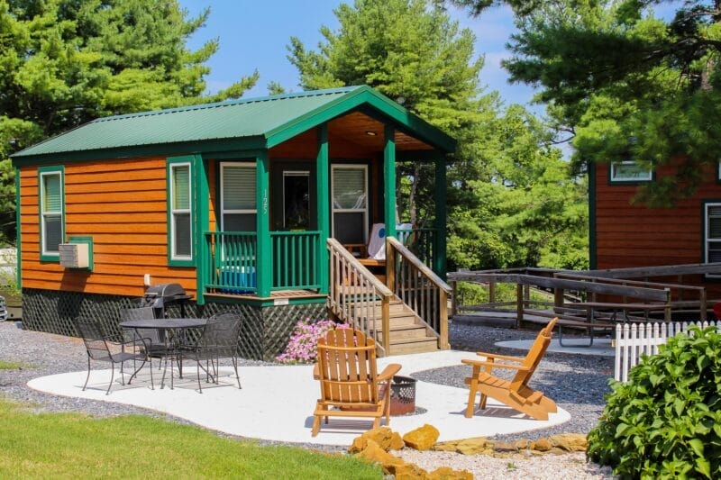 A small wooden cabin with a green roof, outdoor seating area with wooden chairs, a picnic table, and surrounded by trees on a sunny day.