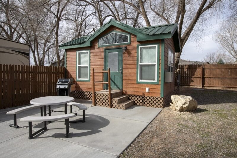 A small wooden cabin with green trim sits next to a picnic table and barbecue grill on a concrete patio, surrounded by a wooden fence and leafless trees.