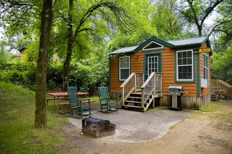 A small wooden cabin with a porch sits among trees. Outside, there is a picnic table, chairs, a grill, and a firepit on a gravel area.