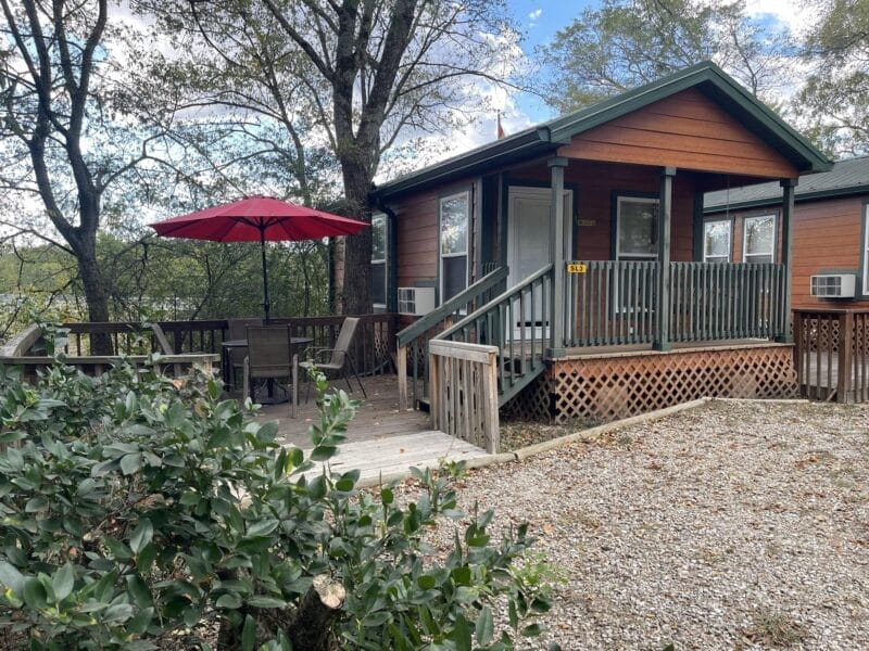 A small wooden cabin with a porch, outdoor seating area, red umbrella, and gravel driveway, surrounded by trees and greenery under a partly cloudy sky.