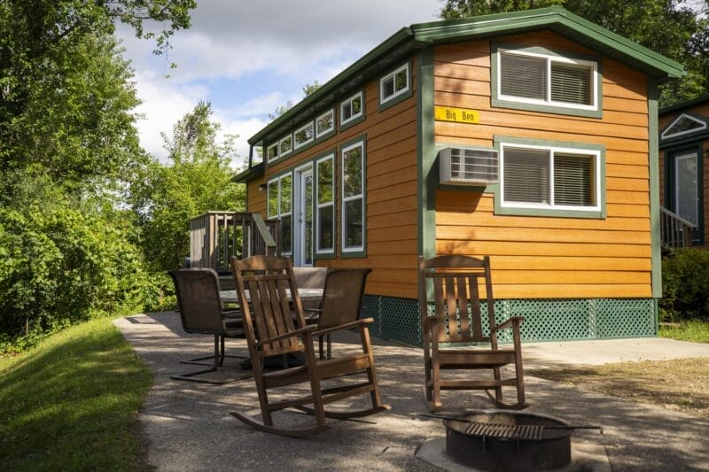 A small orange cabin with green trim, an outdoor patio area with chairs and rocking chairs, and a firepit, surrounded by trees and greenery.