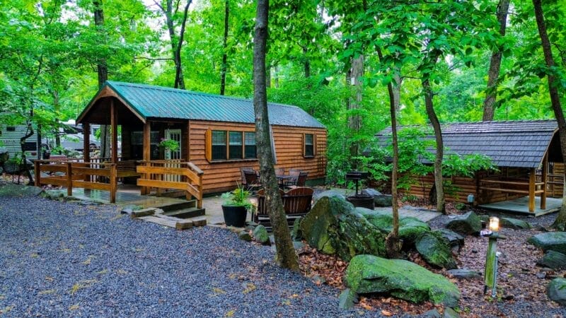A wooden cabin and gazebo sit among trees and large rocks in a forested area, with gravel pathways and outdoor seating visible.