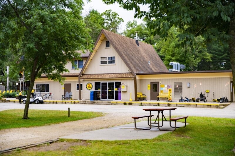 A tan A-frame building with yellow KOA signage, bikes, and karts parked outside, surrounded by trees and a picnic table in the foreground.