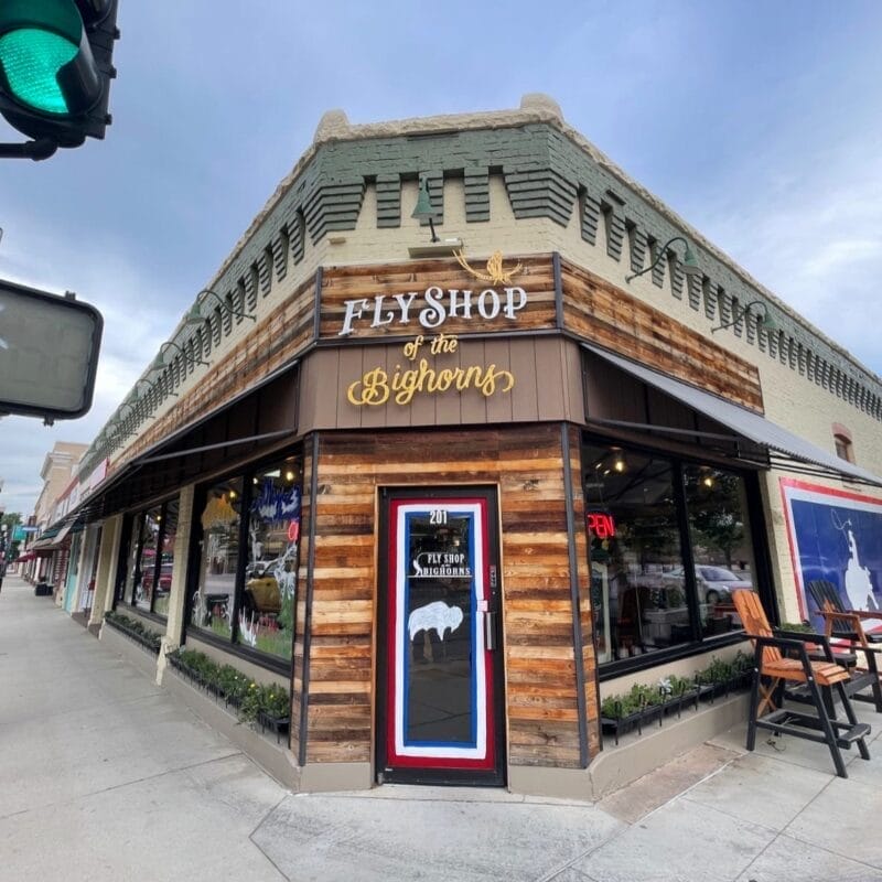 Corner view of "Fly Shop of the Bighorns," a store with wood accents, a sign above the entrance, and fly fishing displays visible through large windows.