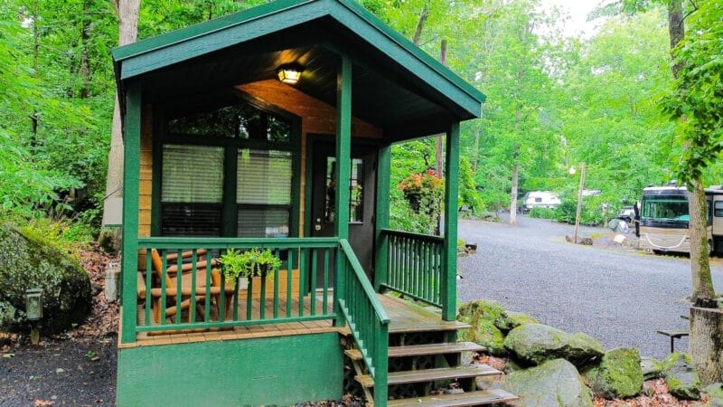 A small green cabin with a covered porch and steps sits among trees near a gravel road, with RVs parked in the background.