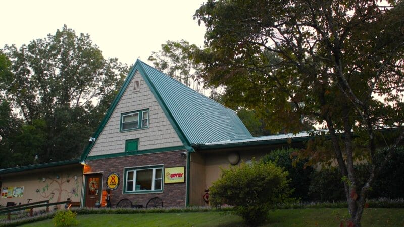 A building with a steep green metal roof, brick and siding exterior, and KOA signage, surrounded by grass, trees, and bushes.