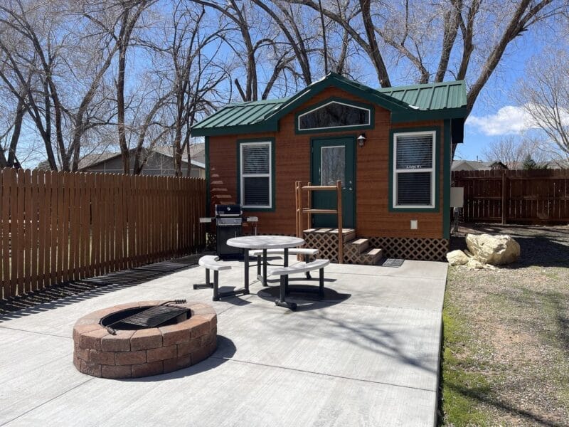 A small wooden cabin with a green roof, a picnic table, a barbecue grill, and a circular fire pit on a concrete patio, surrounded by a wooden fence and leafless trees.