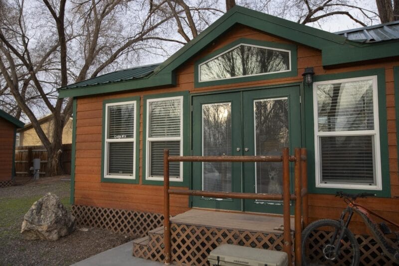 A small wooden cabin with green trim, a front porch with a railing, and a bicycle parked beside it. Leafless trees and another cabin are in the background.