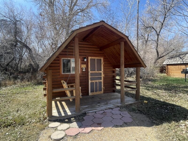 A small wooden cabin with a front porch, swing bench, and the number 2 on the door, surrounded by leafless trees on a sunny day.