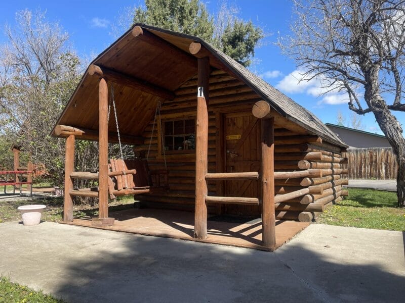 A small log cabin with a covered porch and a wooden swing, surrounded by trees and a concrete walkway under a blue sky.