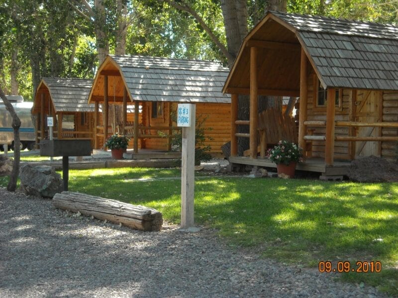 Three small wooden cabins with covered porches are situated on a grassy area with trees and potted flowers nearby. A gravel path runs in front.