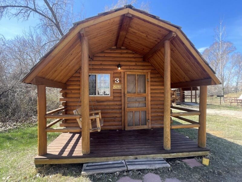 A small wooden cabin with a covered porch, swing chair, and the number 3 on the door, surrounded by grass and trees.