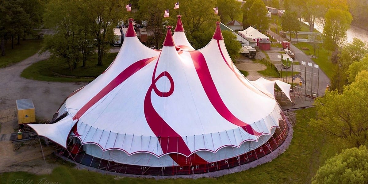 A large white circus tent with red stripes and multiple peaks is set up in a grassy area with surrounding trees and nearby buildings.