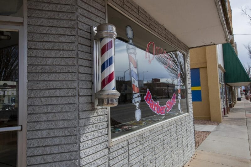 Exterior view of a barber shop with a classic red, white, and blue barber pole and a large window displaying barber-themed graphics and signage.