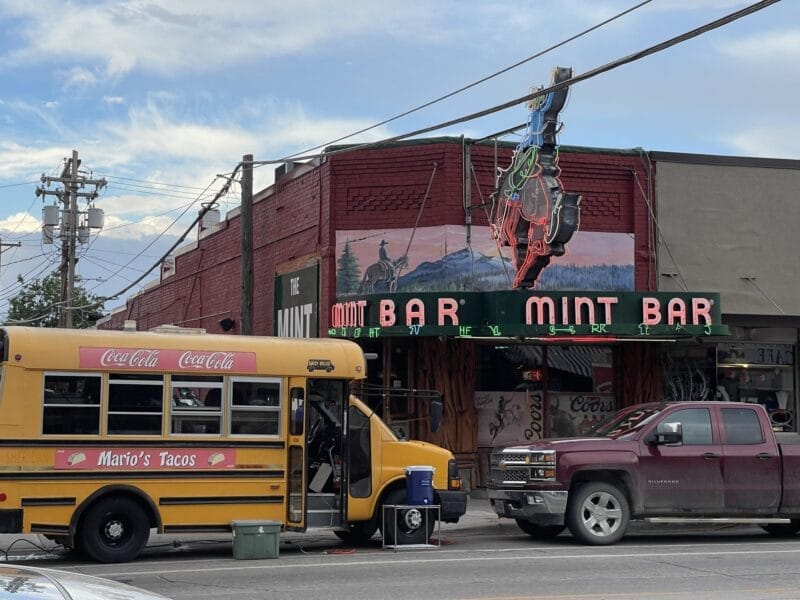 A yellow Mario’s Tacos bus and a maroon pickup truck are parked in front of the Mint Bar, which has a neon cowboy sign and mural on a brick building.