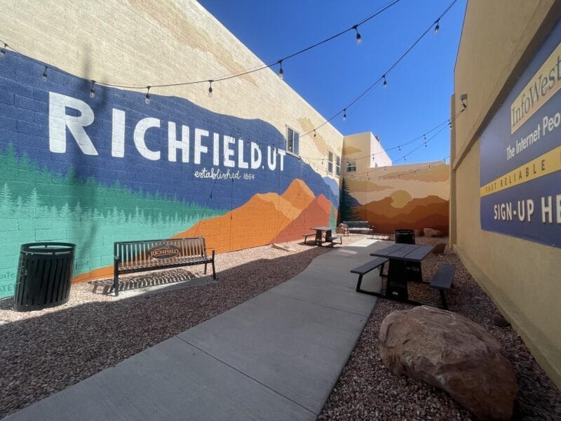 Outdoor seating area with benches and tables next to a painted mural reading "Richfield, UT," surrounded by string lights and advertising signs on the walls.