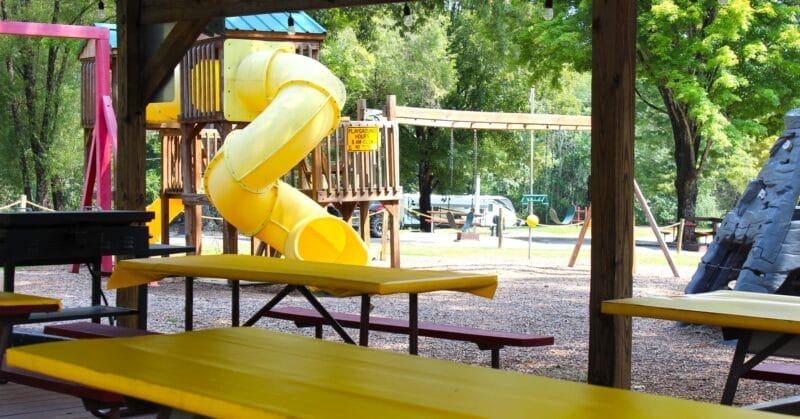 A covered picnic area with yellow tables overlooks a playground featuring a yellow spiral slide, climbing structure, and swings, surrounded by trees on a sunny day.