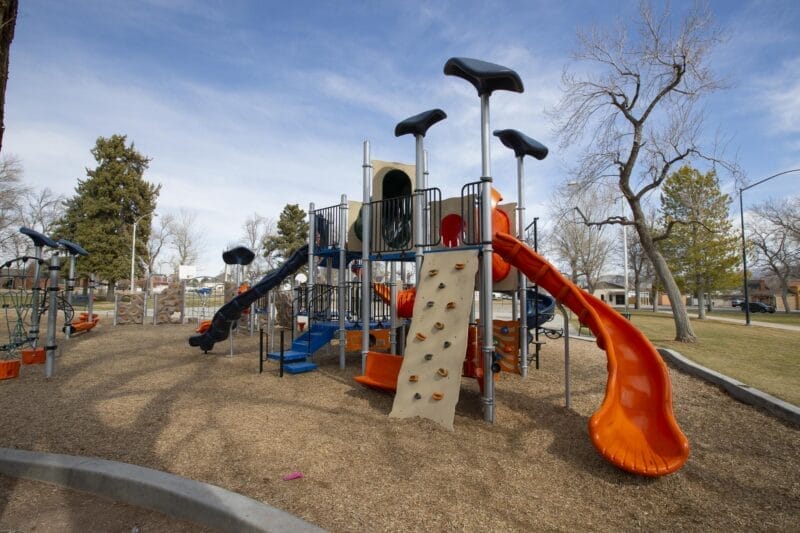 Modern playground structure with multiple slides, climbing walls, and platforms on a wood chip ground, set in a park with trees and grass under a clear sky.