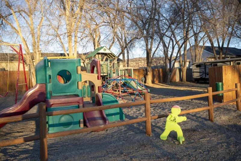 A colorful playground with climbing structures and slides sits behind a wooden fence; a yellow safety figure stands near the fence, with trees and houses in the background.