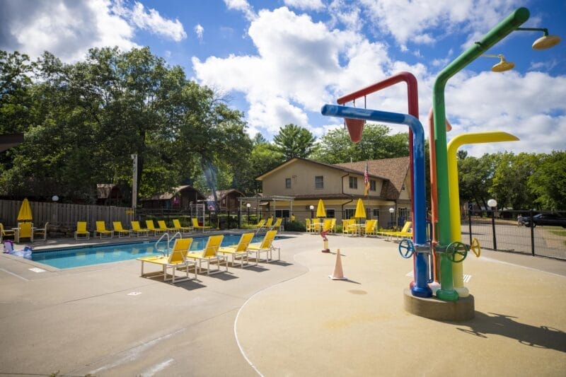 Outdoor pool area with yellow lounge chairs, umbrellas, and a colorful splash pad feature; trees and a building are visible in the background under a partly cloudy sky.