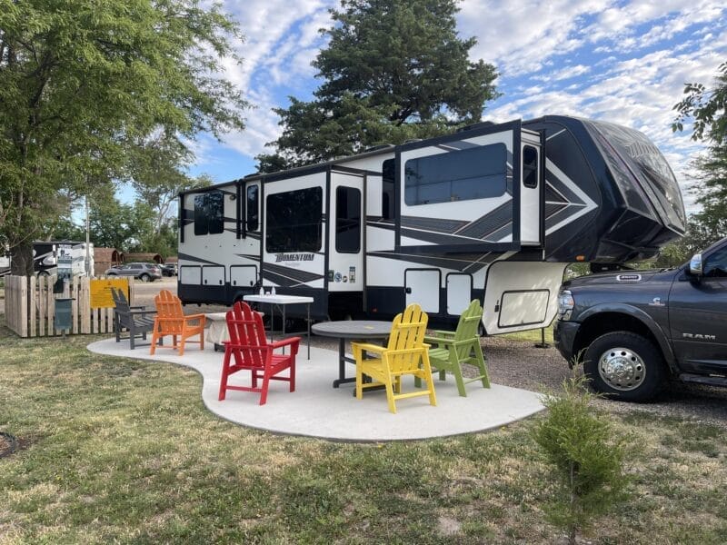 A large RV is parked beside a black pickup truck with five colorful Adirondack chairs and a round table arranged on a concrete patio in front. Trees and grass surround the area.