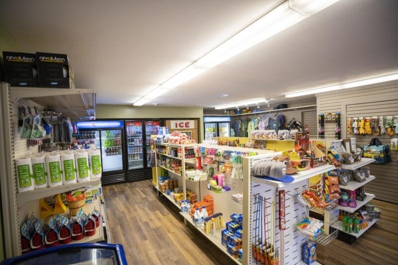 A small convenience store interior with shelves stocked with snacks, drinks, toiletries, pet supplies, and refrigerators against the back wall.