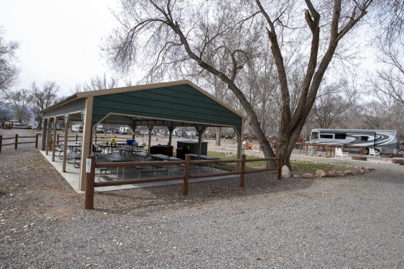 A covered picnic area with tables and benches is surrounded by leafless trees and a gravel path; an RV is parked in the background.