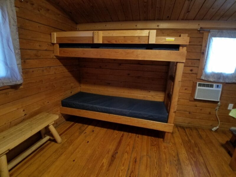 Wooden bunk bed with dark blue mattresses in a small cabin room with wooden walls and floor, a bench, two windows with curtains, and a wall-mounted air conditioner.