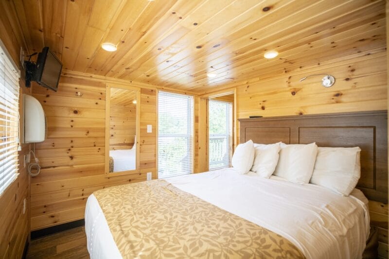 A small bedroom with wooden walls and ceiling, a bed with white linens and pillows, a wall-mounted TV, a mirror, and large windows with blinds.